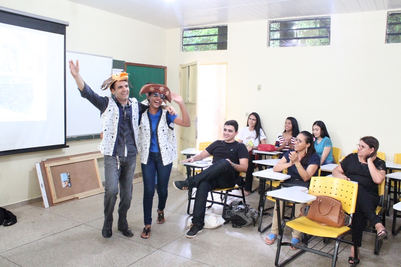 Duas pessoas com chapéu na cabeça e de pé cantando enquanto as demais os observam sentados na sala de aula 