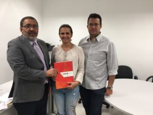 Três pessoas de pé segurando um documento e sorrindo para a pessoa que faz a fotografia destas