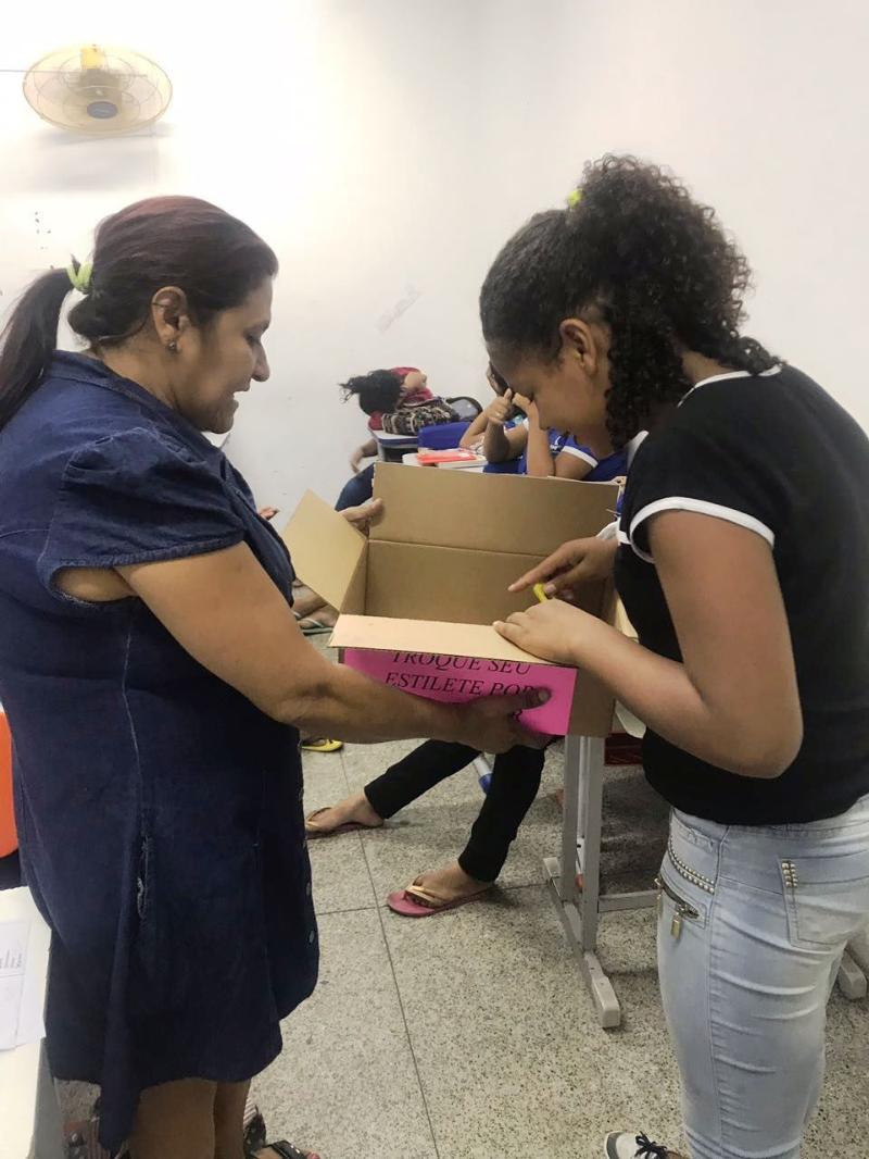 Duas pessoas de pé segurando uma caixa. Uma das pessoas coloca objetos dentro da caixa 