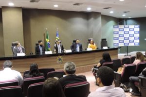 Pessoas sentadas em auditório. Ao fundo as pessoas observam os chefes de insituições que estão assentados a sua frente