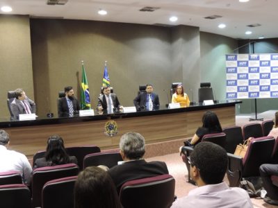 Pessoas sentadas em auditório. Ao fundo as pessoas observam os chefes de insituições que estão assentados a sua frente