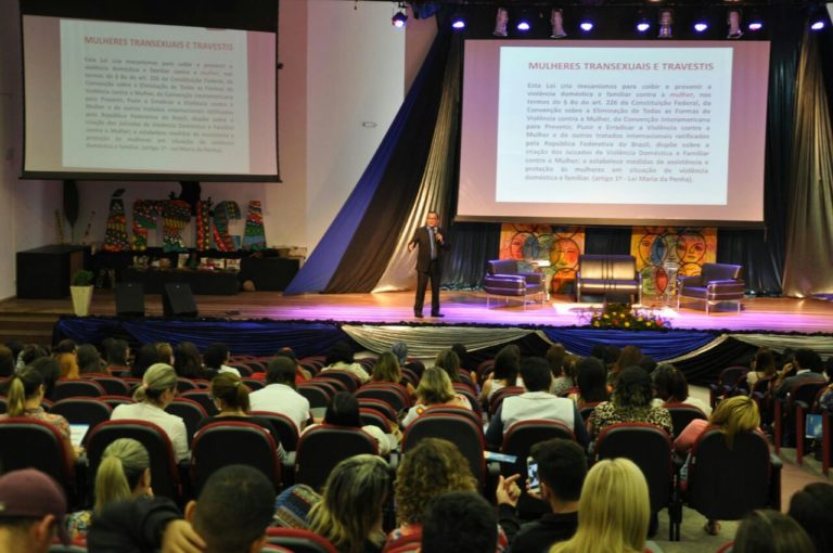 Pessoas sentadas em auditório ao fundo um homem de pé no palco falando com microfone nas mãos