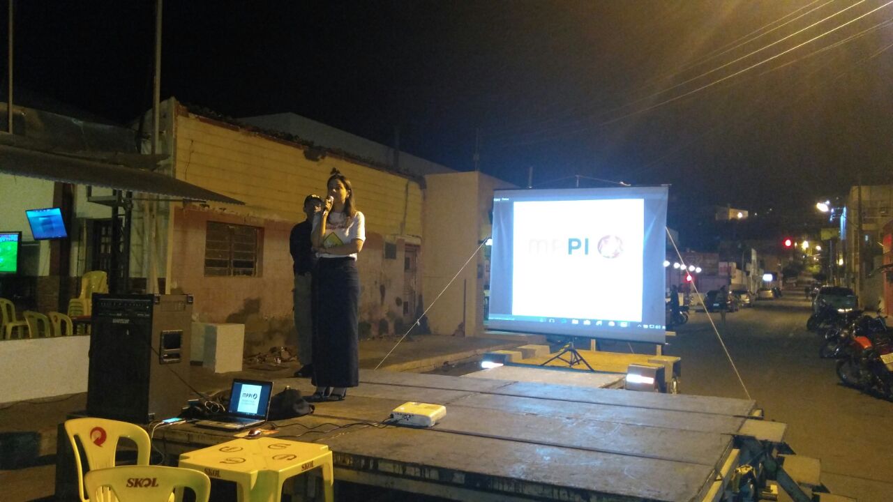 Pessoas de pé em um rua de Picos. Na rua há um palco com uma mulher e um homem falando sobre trânsito 