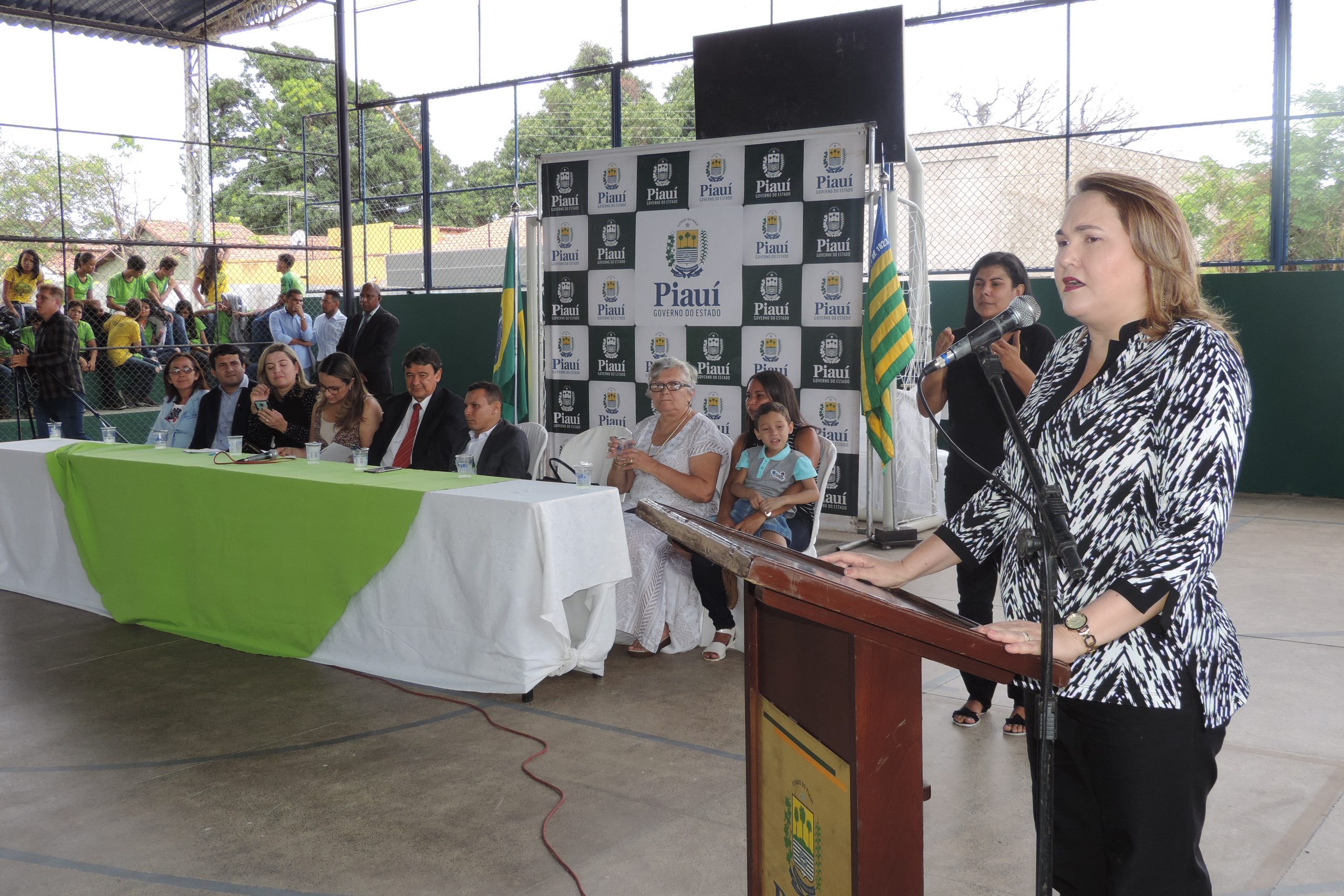 Mulher de pé falando ao fundo pessoas sentadas em uma mesa a observam 