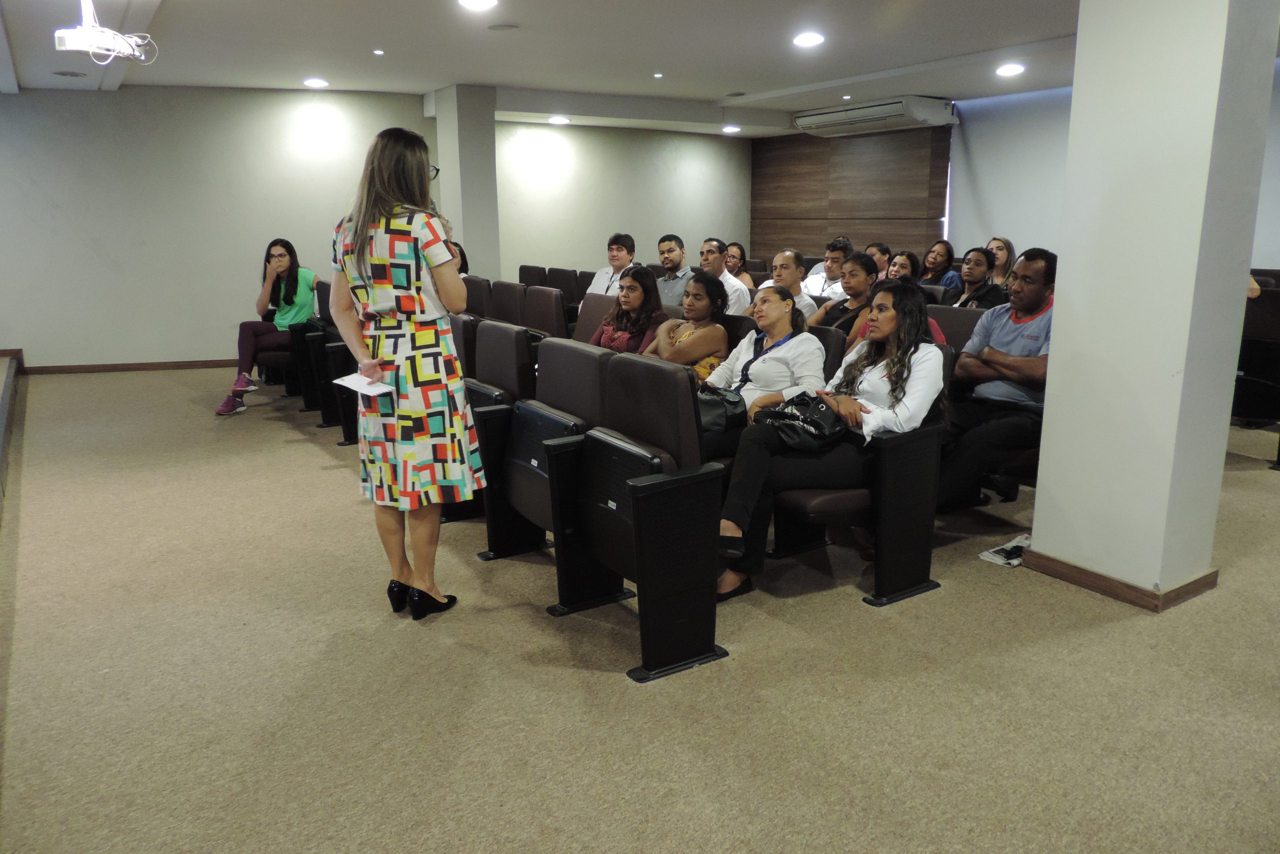 Mulher de pé com microfone em mãos conversando com pessoas que estão sentadas a sua frente 
