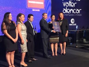 Pessoas de pé num palco. Ao centro três pessoas seguram uma placa do prêmio