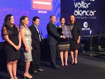 Pessoas de pé num palco. Ao centro três pessoas seguram uma placa do prêmio