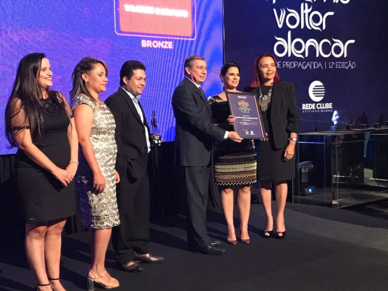 Pessoas de pé num palco. Ao centro três pessoas seguram uma placa do prêmio