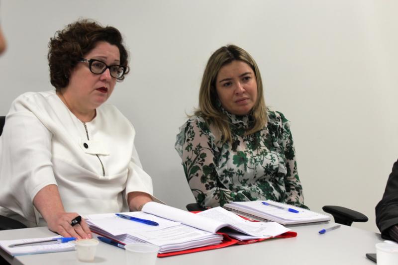 Duas pessoas sentadas em volta de uma mesa observando outras pessoas que falam no momento 