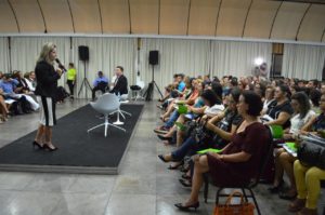 Uma mulher de pé falando com microfone nas mãos olhando para pessoas que estão sentadas à sua frente