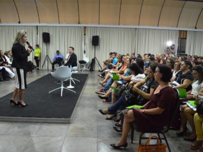 Uma mulher de pé falando com microfone nas mãos olhando para pessoas que estão sentadas à sua frente