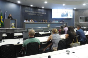 Pessoas sentadas olhando para uma mulher que está de pé falando da tribuna
