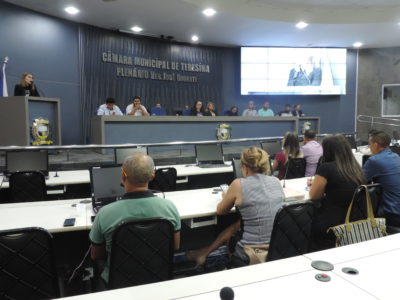 Pessoas sentadas olhando para uma mulher que está de pé falando da tribuna