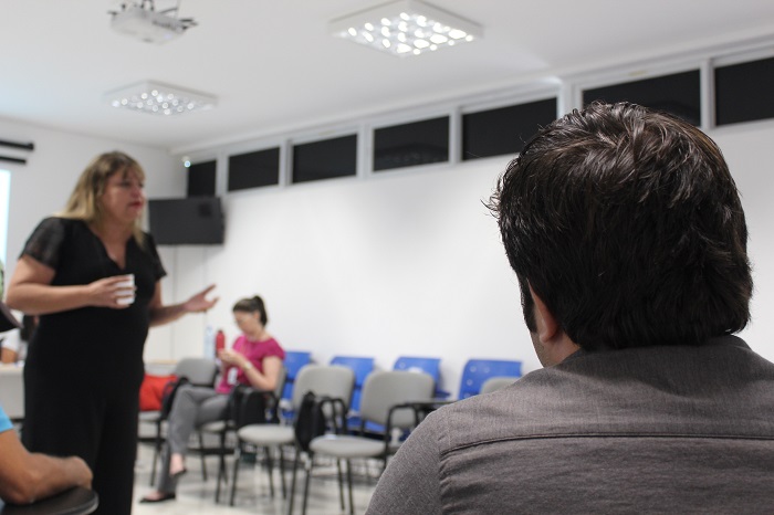 Homem sentado em uma cadeira e ao fundo uma mulher de pé falando 