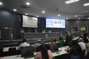 Pessoas sentadas observando mulher que fala da tribuna