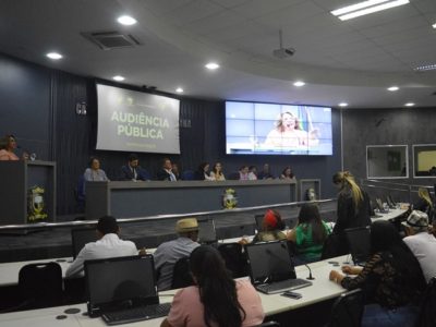 Pessoas sentadas observando mulher que fala da tribuna
