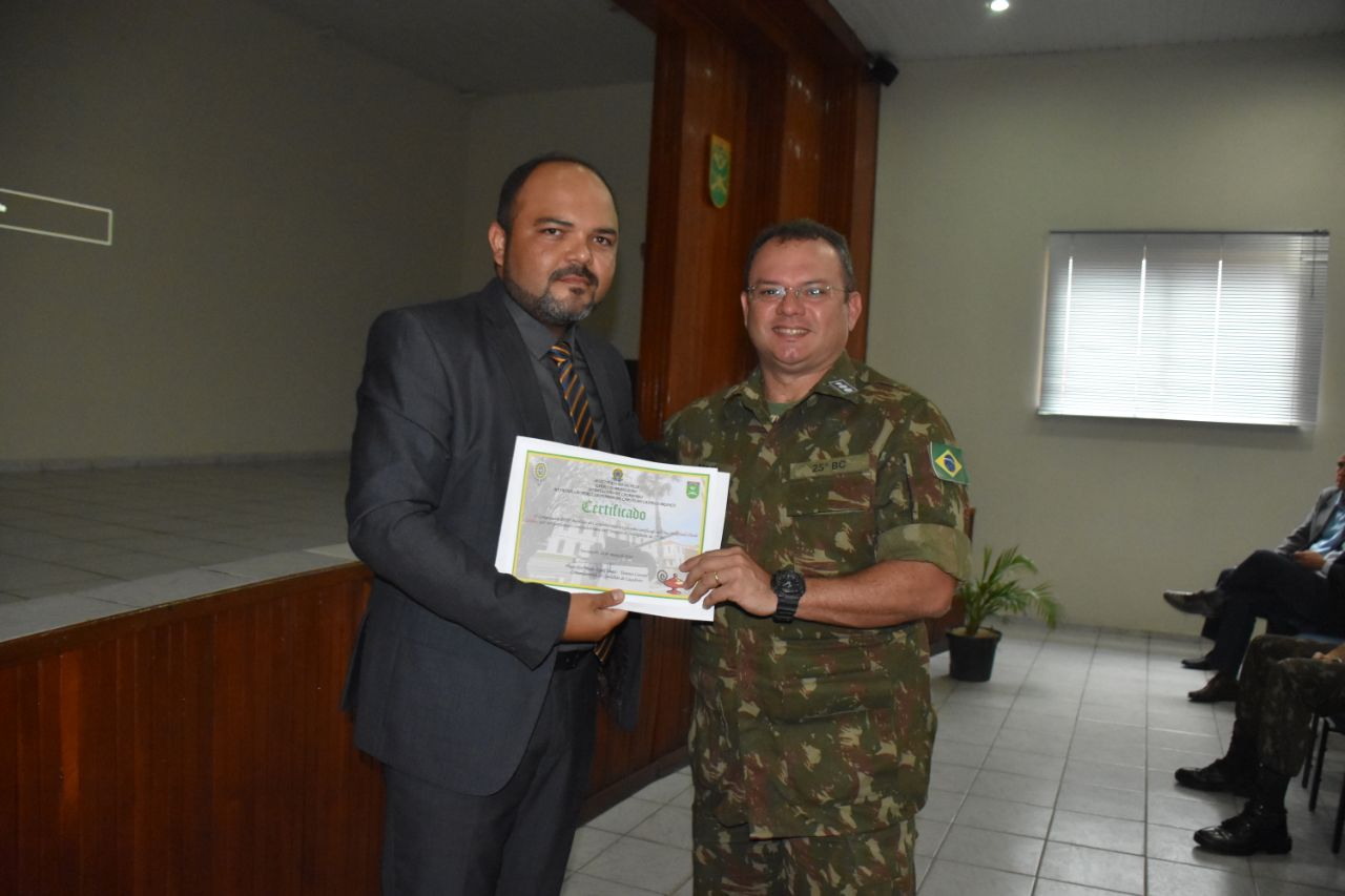 Duas pessoas de pé. Um entrega um diploma para o outro. 