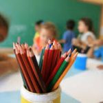 Foto que mostra, em primeiro plano, vários lápis coloridos. Ao fundo, crianças desenham na escola.