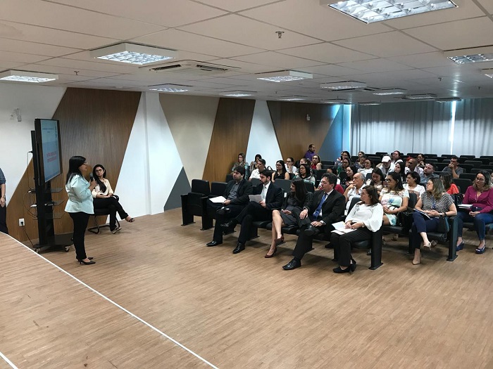 Pessoas sentadas em auditório vendo mulher de pé falar 