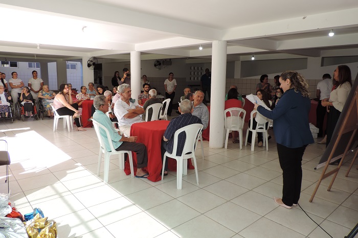 Pessoas sentadas em um grande salão. No momento uma mulher de pé fala aos presentes que a observam. 