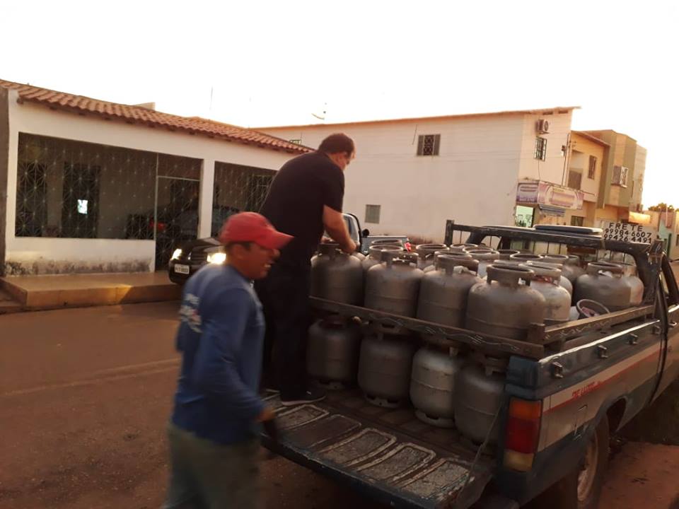 Dois homens atrás de um veículo. Um deste coloca um botijão na carroceria do carro 