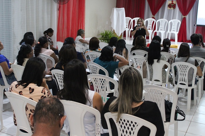 Pessoas sentadas em auditório, observando mulher que está de pé falando para plateia