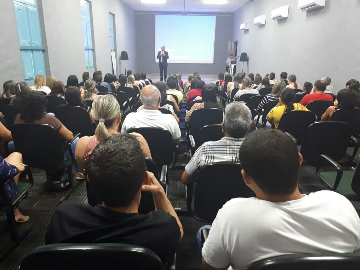 Pessoas sentadas em auditório observando um homem que está de pé no palco falando 