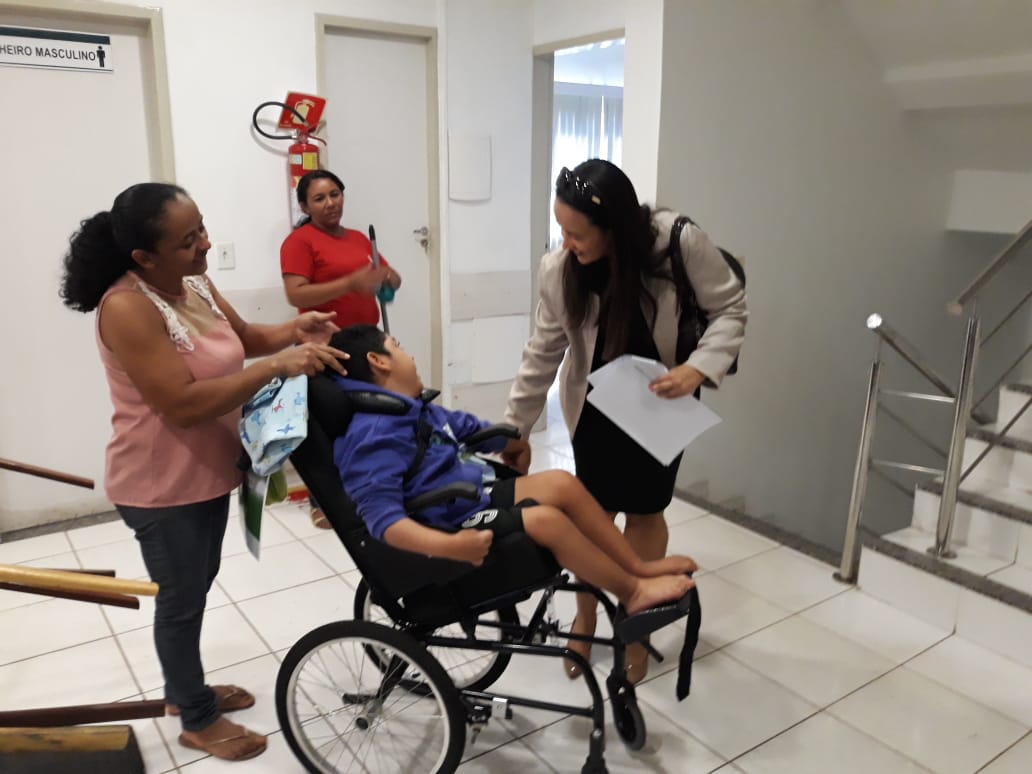 Pessoas conversando entre si. Uma mulher está de pé e cumprimenta uma senhora cadeirante 