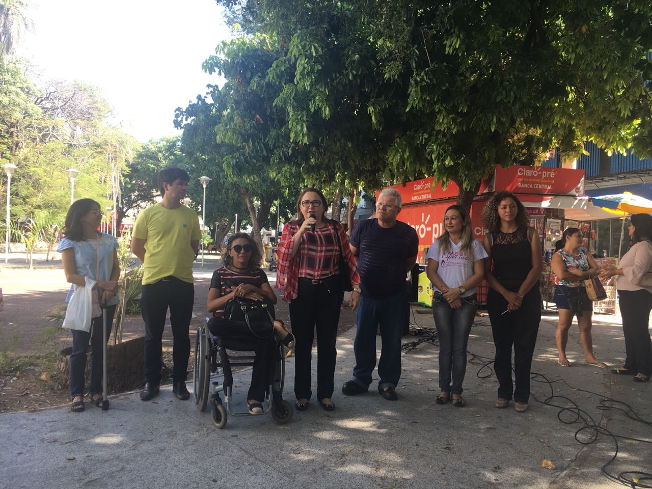 Pessoas de pé. AO centro há uma mulher com microfone na mão, falando para o público que a observa 