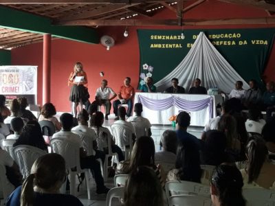 Pessoas sentadas em um auditório, observando uma mulher que está de pé falando para estes