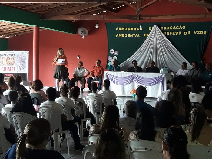 Pessoas sentadas em um auditório, observando uma mulher que está de pé falando para estes