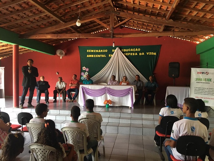 Pessoas sentadas em auditório observando um homem que está de pé no palco falando 