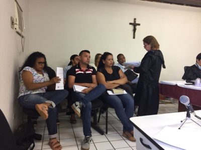 Pessoas sentadas observando um mulher que está de pé falando com documentos nas mãos