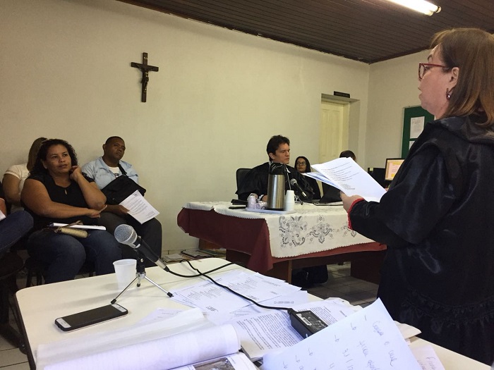 Pessoas sentadas observando um mulher que está de pé falando com documentos nas mãos 