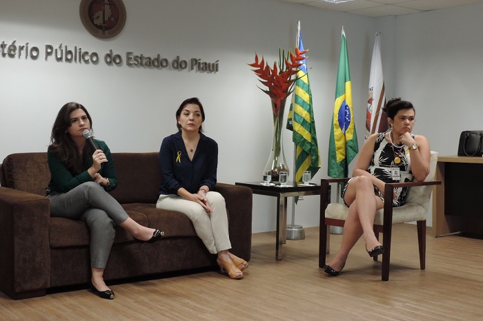 Três mulheres entadas em um semicírculo. Uma delas com microfone nas mãos 