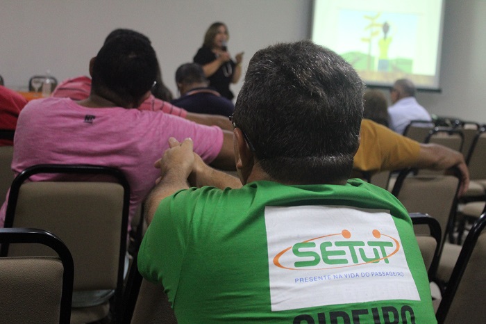 Homem sentando olhando para a promotora de Justiça Amparo Paz que está ao fundo proferindo palestra sobre violência doméstica 