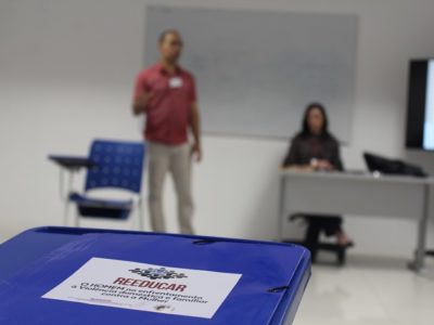 Foto de uma pasta sobre uma cadeira com o nome do projeto Reeducar. Ao fundo há um homem de pé falando para a plateia