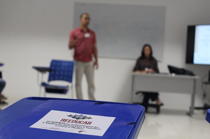 Foto de uma pasta sobre uma cadeira com o nome do projeto Reeducar. Ao fundo há um homem de pé falando para a plateia