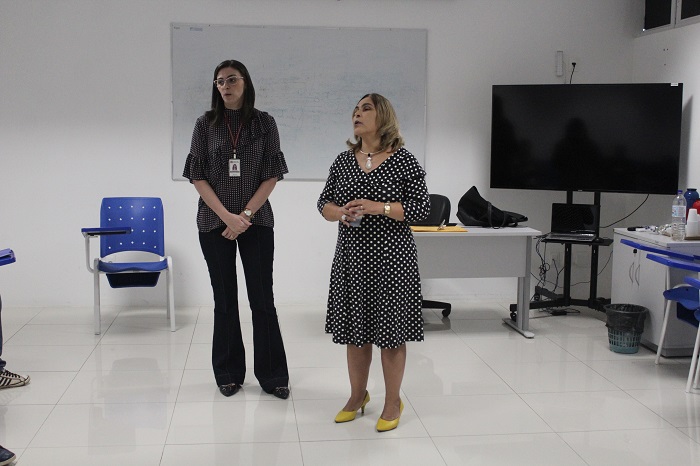Duas mulheres de pé falando com pessoas que estão sentadas a sua frente 