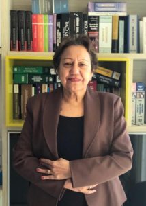 Mulher de pé com os braços cruzado e sorrindo. Atrás há uma estante de livros