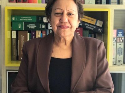 Mulher de pé com os braços cruzado e sorrindo. Atrás há uma estante de livros