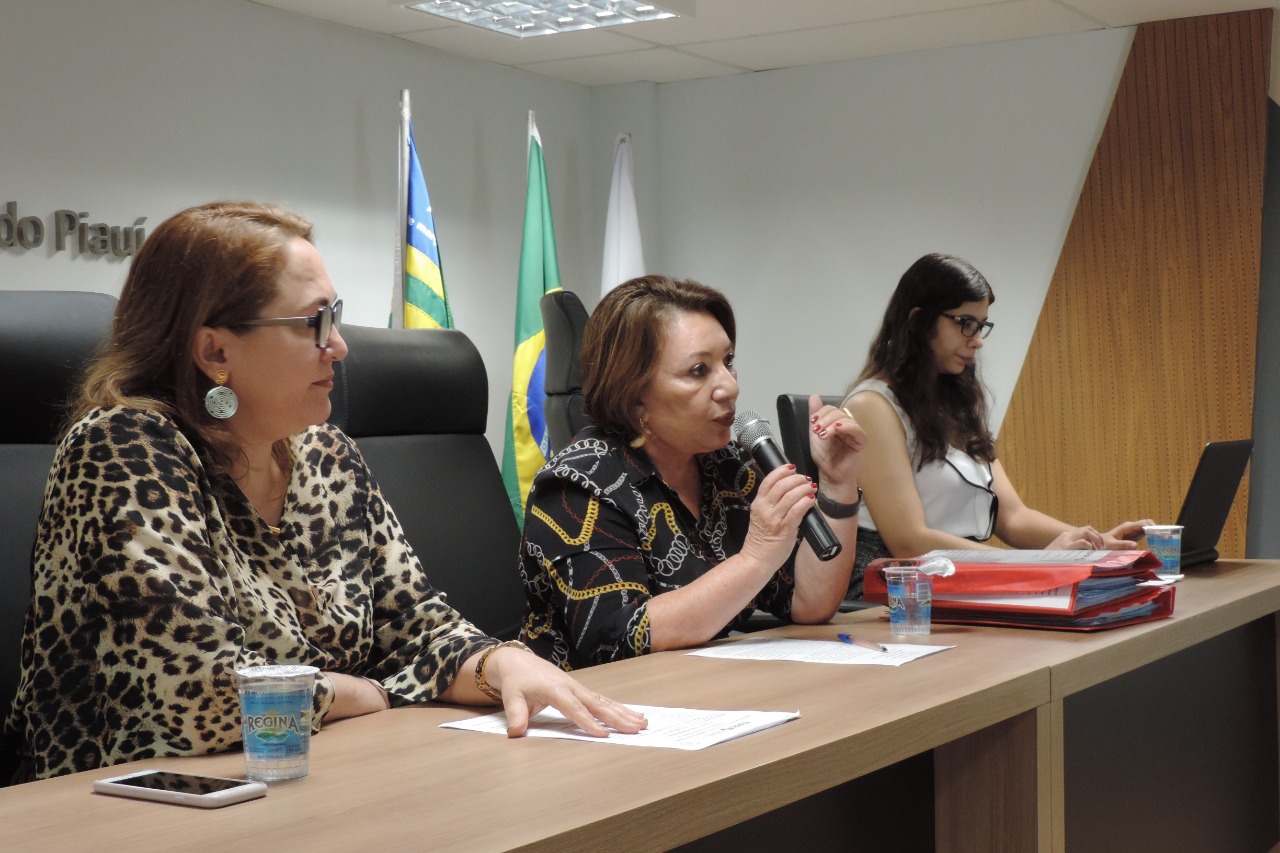 Três mulheres sentadas em uma bancada. Uma das mulheres segura um micorfone nas mãos 