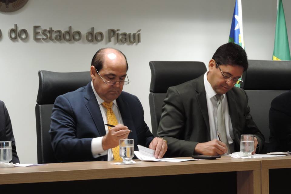 Duas pessoas sentadas assinando documento que está sobre a mesa 