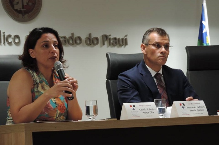 Duas pessoas sentadas em uma mesa. Um dessas pessoas segura um microfone quanto fala com o público que a assiste
