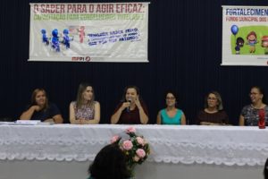 Pessoas sentadas atrás de uma mesa. Uma das pessoas segura um microfone nas mãos