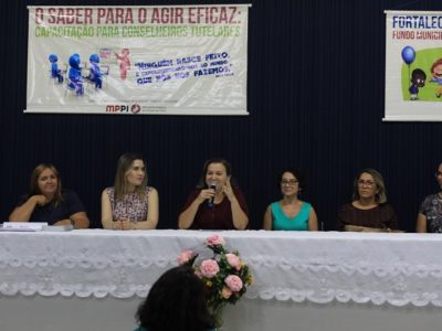 Pessoas sentadas atrás de uma mesa. Uma das pessoas segura um microfone nas mãos