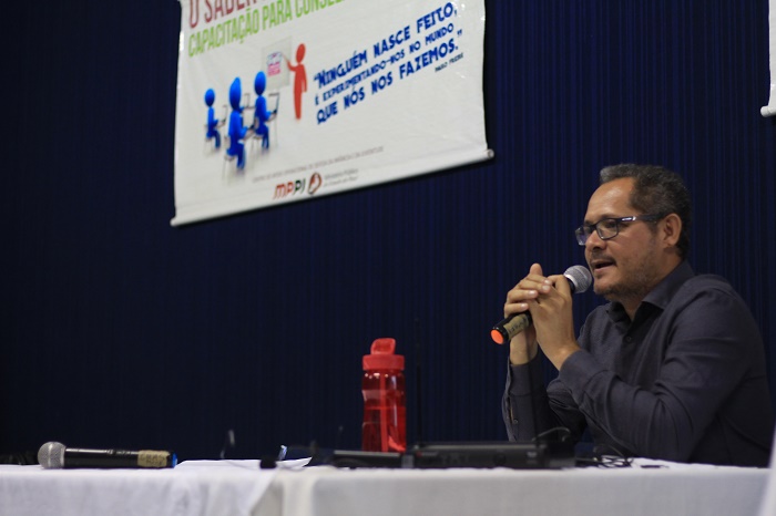 Homem sentado atrás de uma mesa falando com microfone nas mãos 