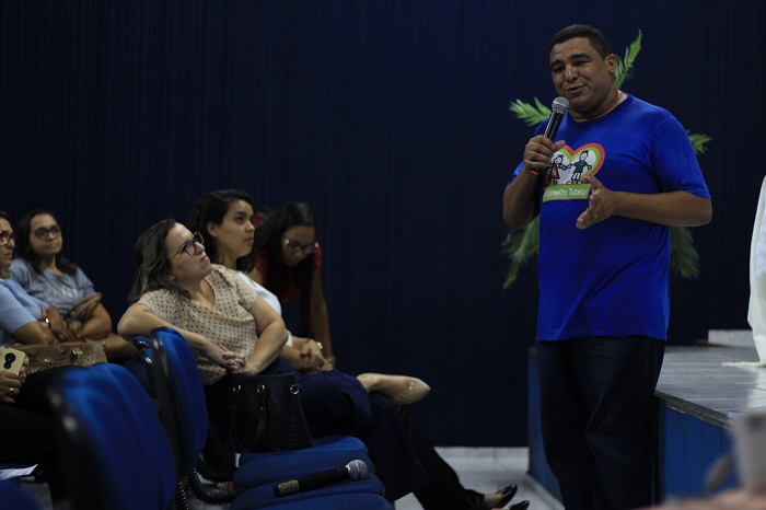 Homem de pé falando com um microfone nas mãos. Duas mulheres estão assentadas a sua frente observando enquanto o mesmo fala 