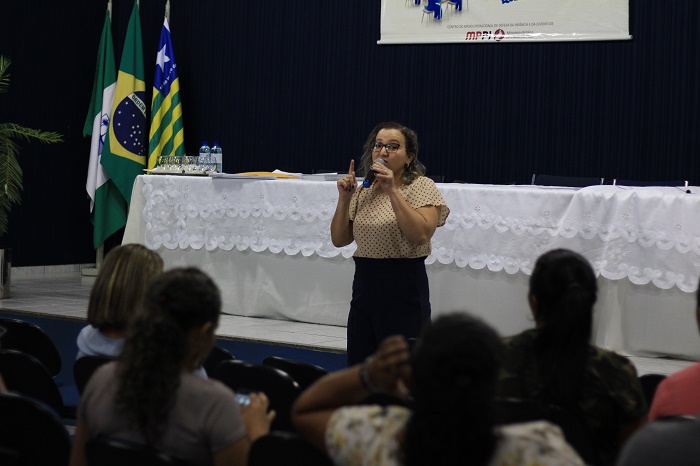 Mulher de pé com microfone nas mãos falando para plateia 
