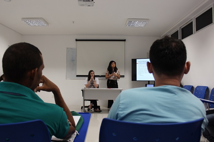 Ao fundo duas mulheres de pé falando com homens que estão na plateia 